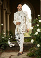 Man wearing a traditional white embroidered Prince jacket and pants standing in a garden with flowers and a stone archway.