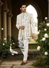 Man wearing a traditional white embroidered Prince jacket and pants standing in a garden with flowers and a stone archway.