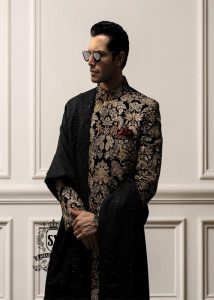 This luxurious royal black velvet sherwani, adorned with opulent gold hand embroidery, exudes a bold and dignified presence. The plush velvet fabric highlights the depth of the intricate hand embellishments, creating a truly regal style befitting a groom or a grand ceremony.